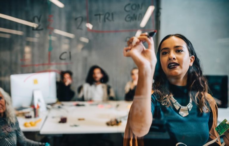 woman writing with marker on a clear glass brainstorming board with group of people at a table behind her