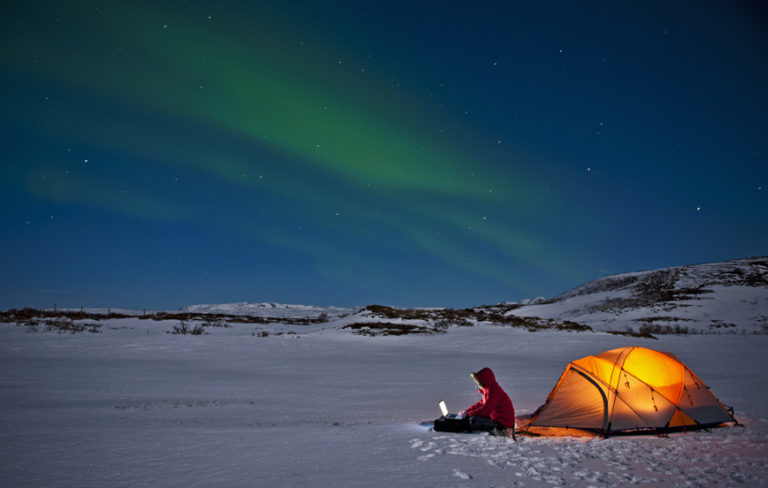 lone person sitting outside lit tent on snow in deserted snowy landscape with norther lights in the sky representing remote work and human connections