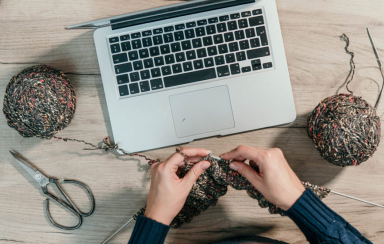 person knitting yarn in front of laptop computer